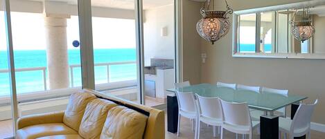 A modern living room with a glass table and white chairs, a yellow leather sofa, and a view of the ocean.