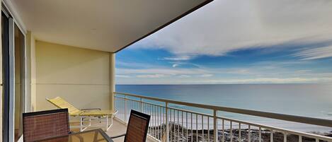 A balcony with a glass table and chairs, overlooking a rocky beach and the ocean.