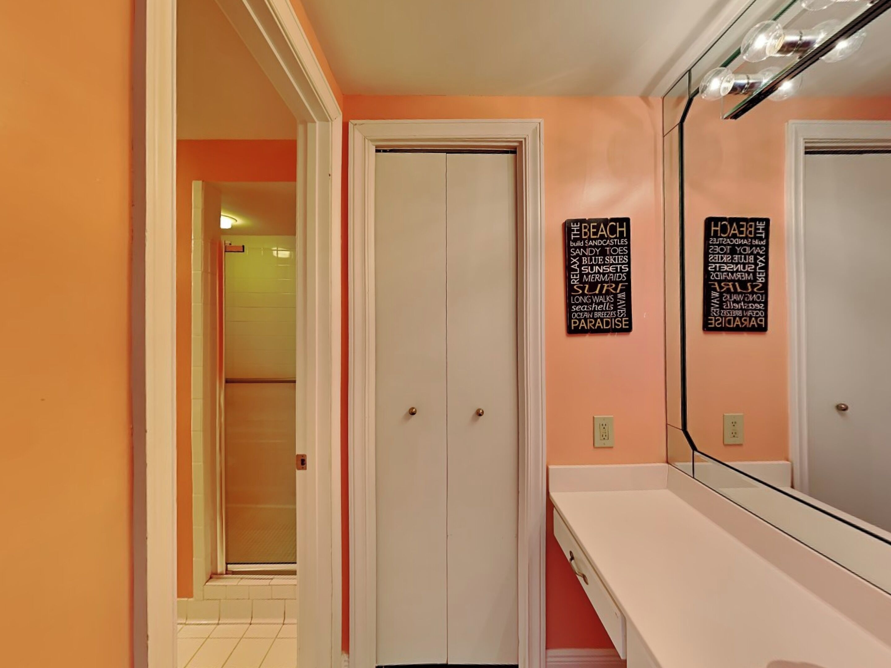 A bathroom with a white vanity, a large mirror, and two doors. There are two signs on the wall with words like "beach," "sandcastles," and "mermaids."