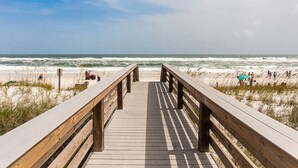 Ein Holzsteg führt zu einem Strand, im Hintergrund sind Wellen zu sehen.
