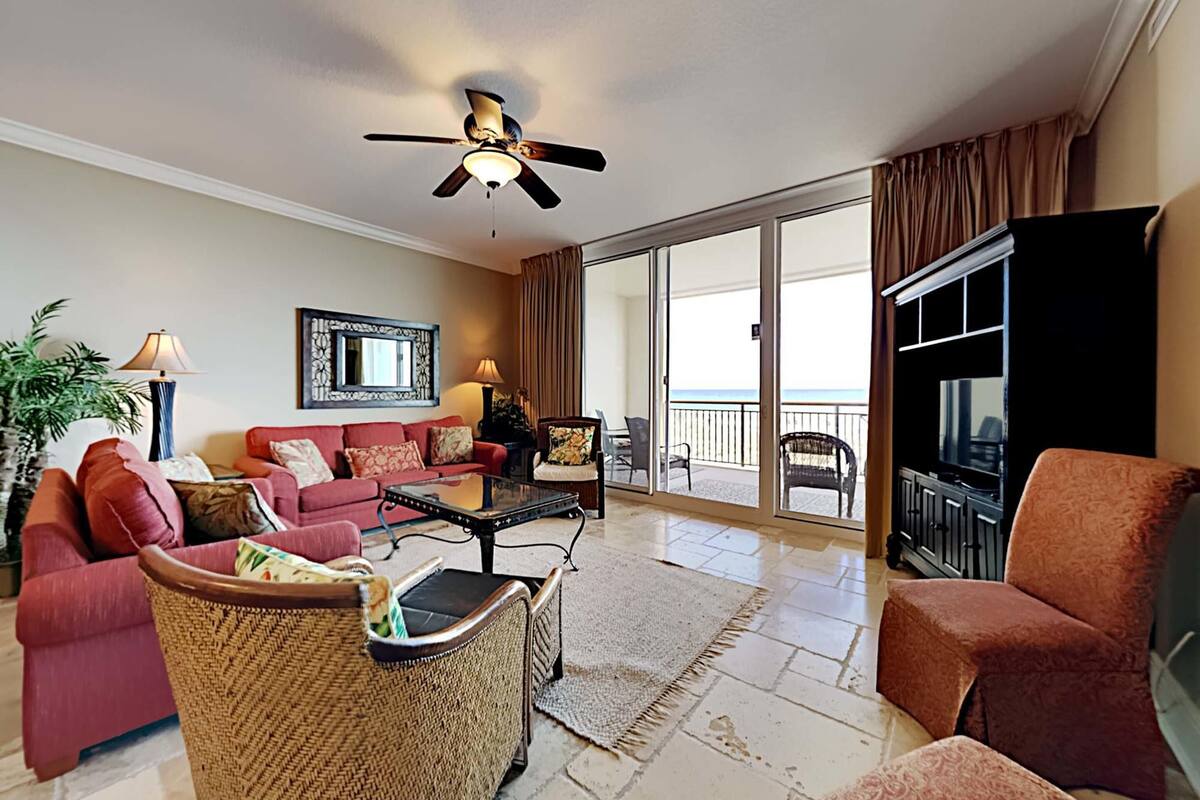 A spacious living room with a red sofa, a wicker chair, a coffee table, and a ceiling fan. A large TV is mounted on the wall, and the room has a view of the ocean through the sliding glass doors.