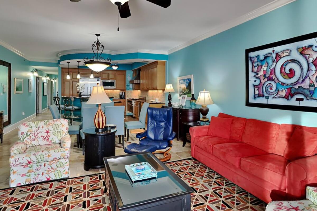 A living room with a red sofa, a blue chair, a floral armchair, a coffee table, and a ceiling fan.
