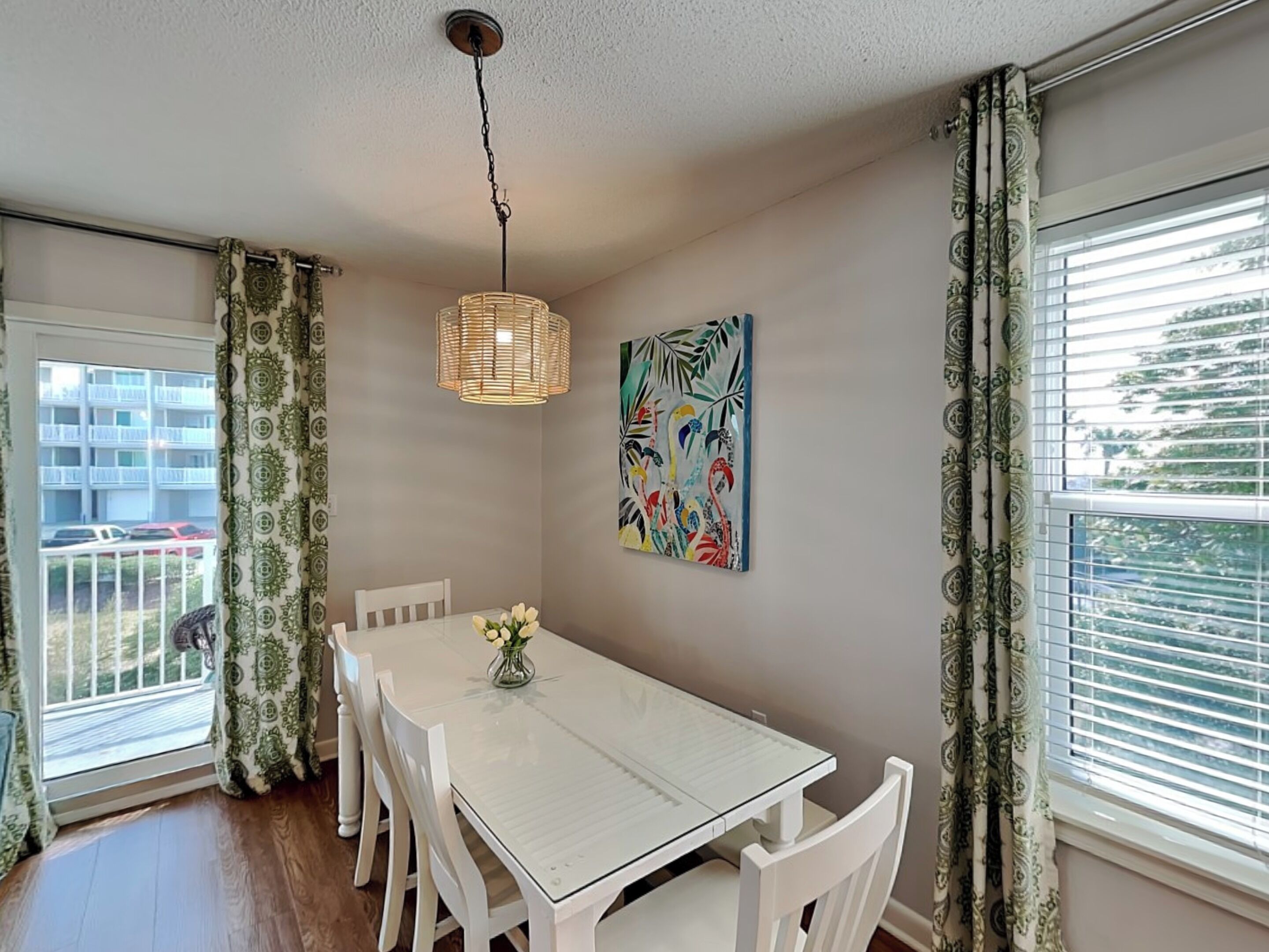 A dining area with a white table and chairs, a hanging light fixture, and a colorful painting on the wall.