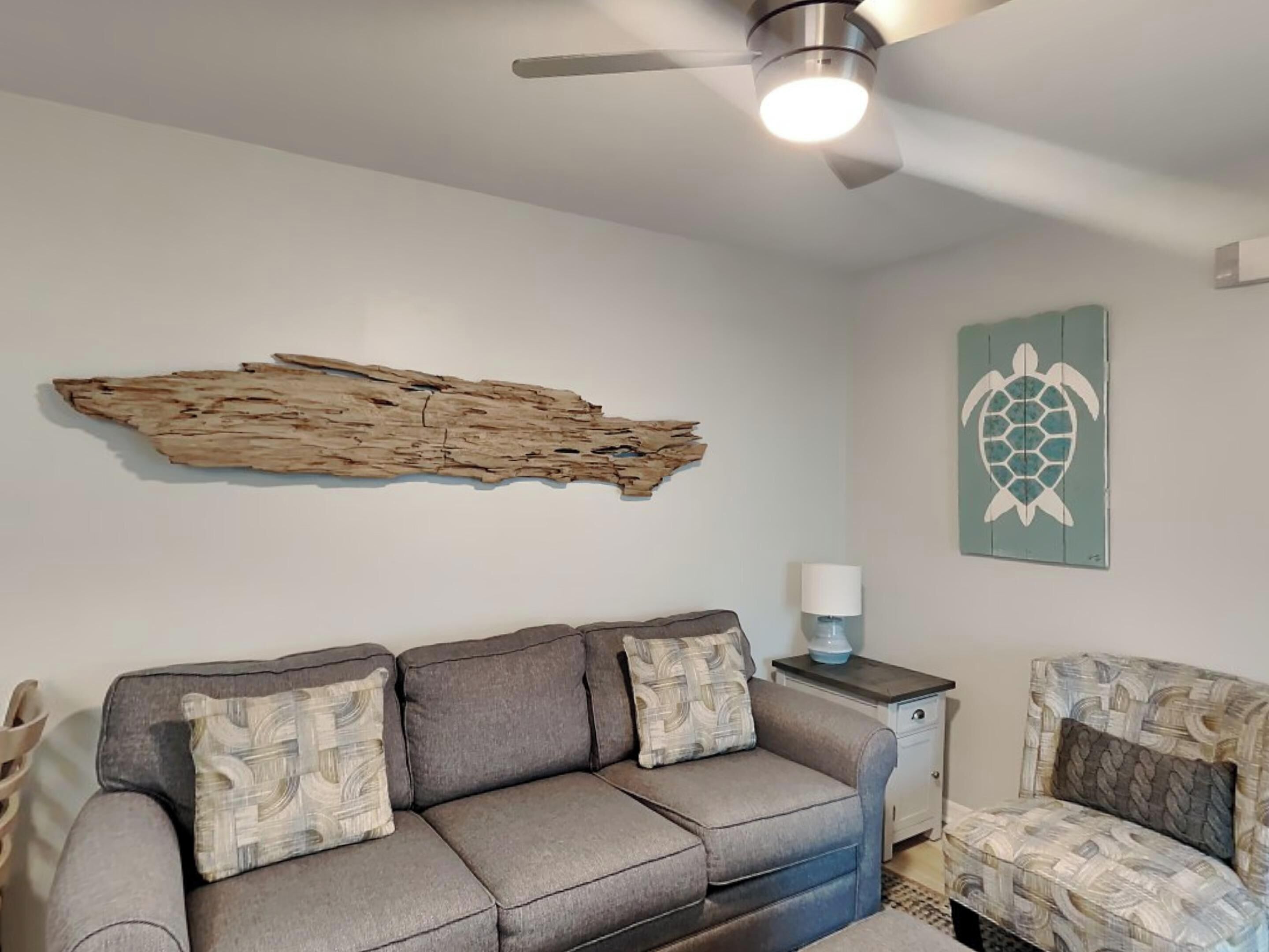 A living room with a gray sofa, a wooden wall art piece, a turtle-themed wall decoration, and a ceiling fan.