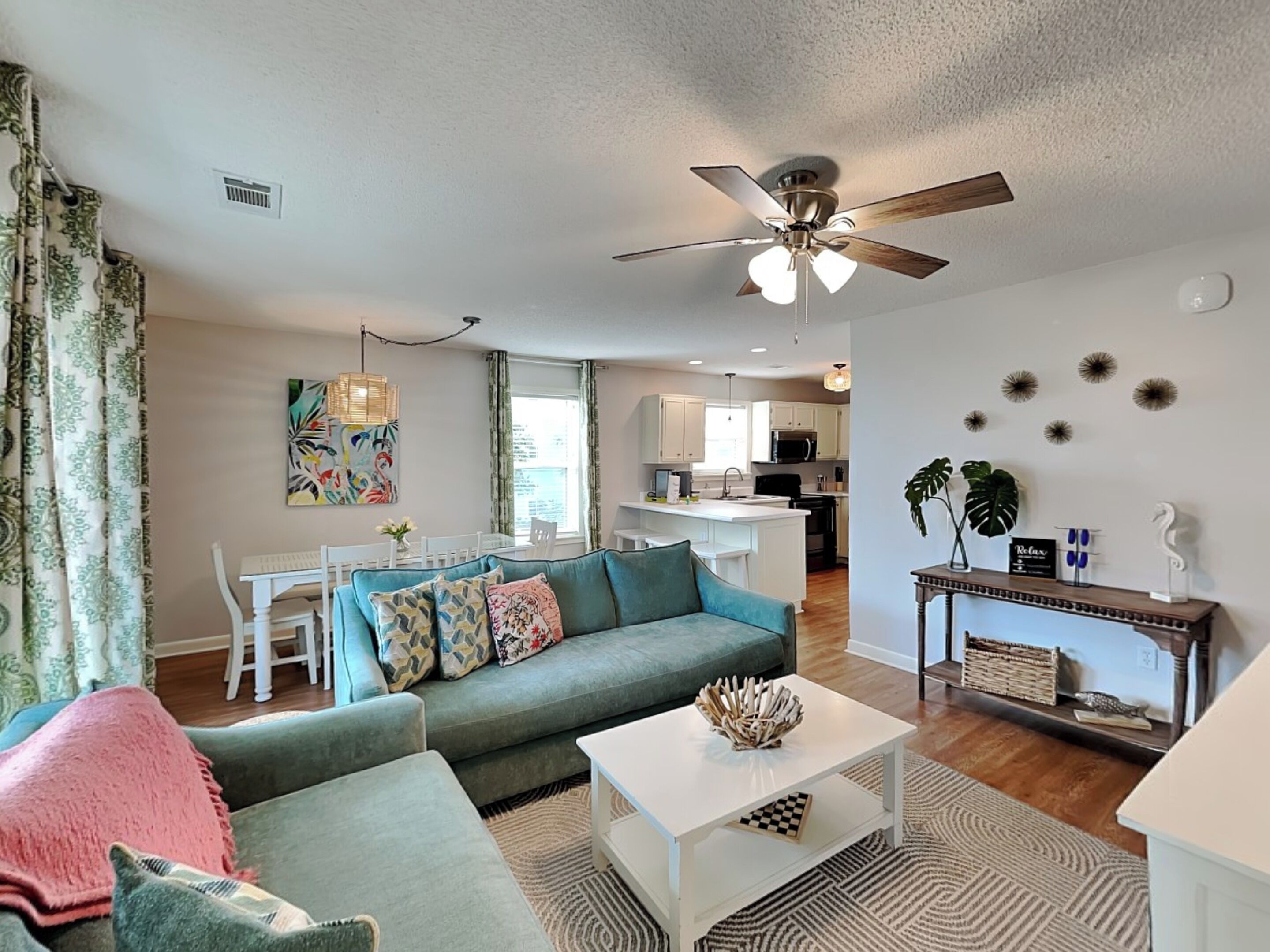 A living room with a sofa, coffee table, and a ceiling fan. There is a dining area with a table and chairs, and a kitchen in the background.