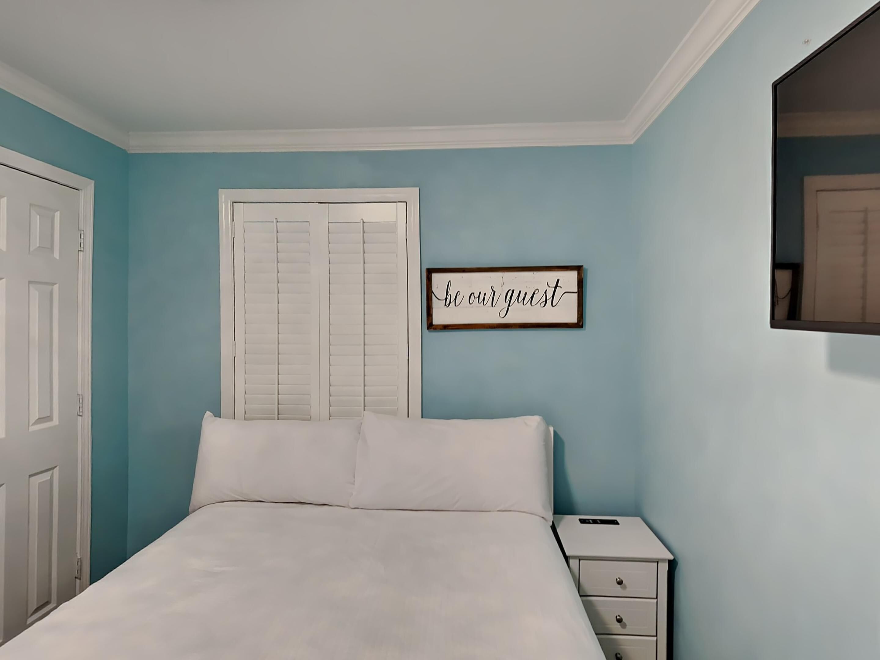 Una camera da letto con un letto, un comodino, un cartello incorniciato e una porta.