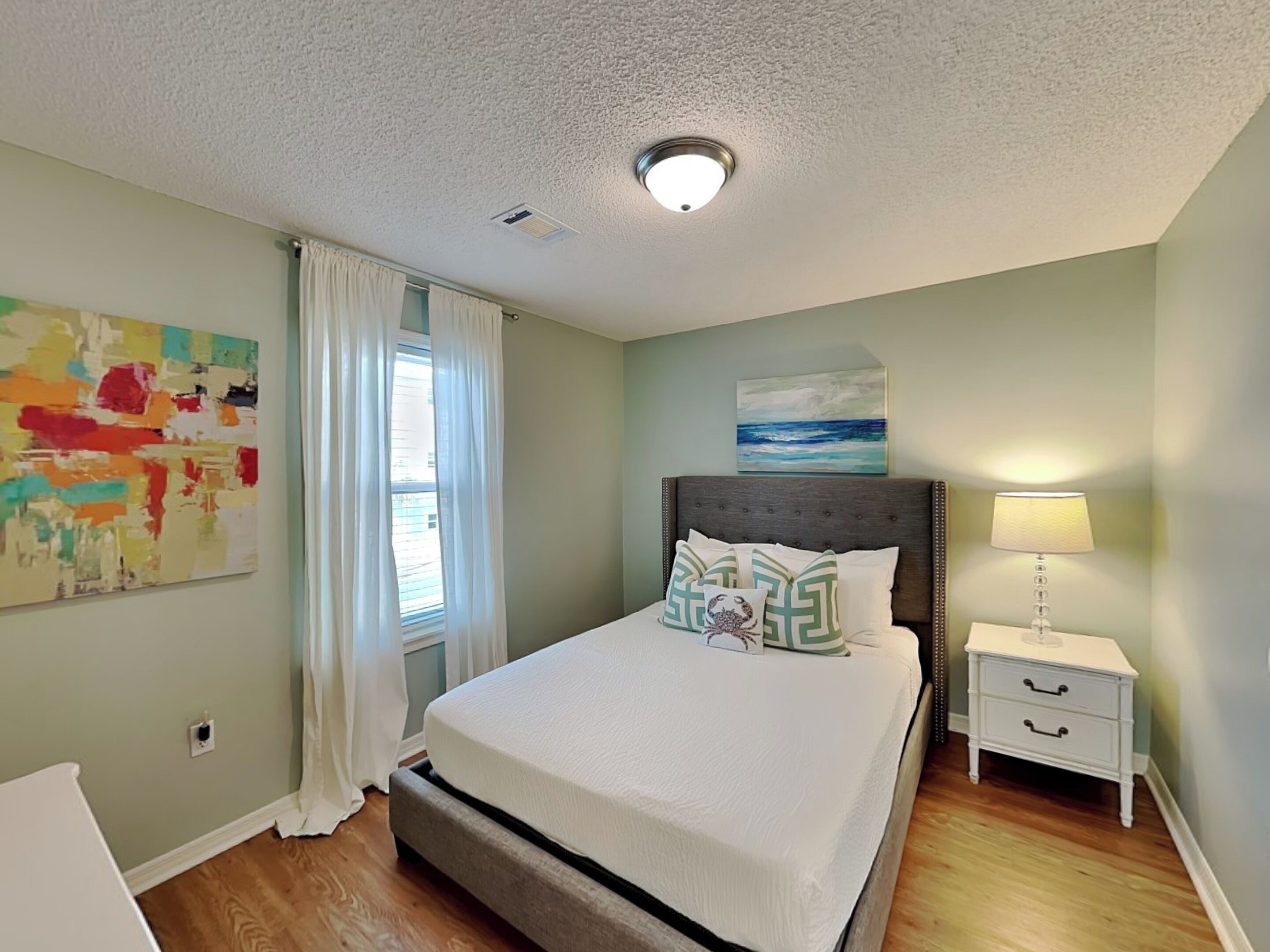 A bedroom with a bed, a nightstand, a lamp, a window with curtains, and a painting on the wall.