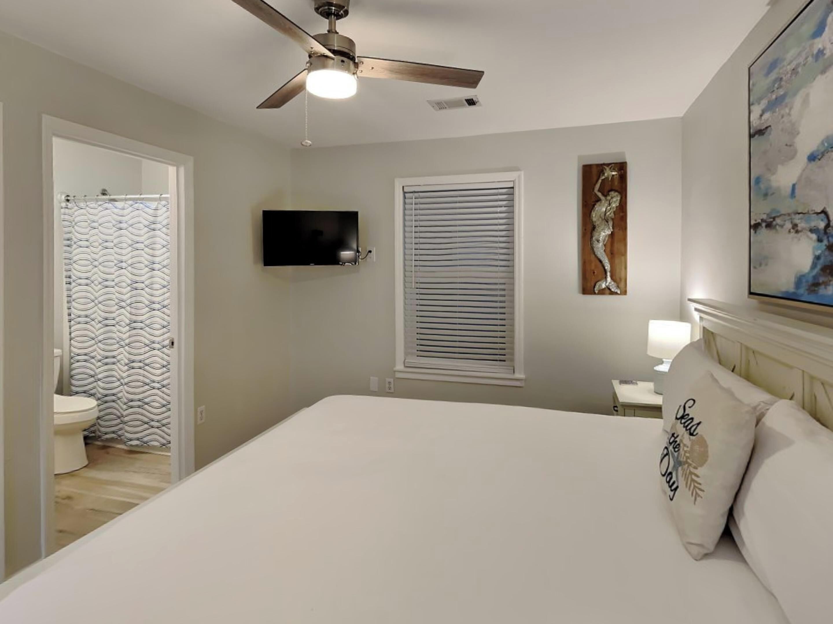 A bedroom with a bed, a ceiling fan, a TV, a window with blinds, a painting, and a nightstand with a lamp.