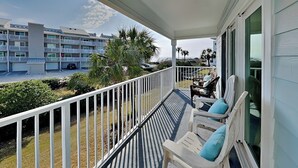 Un balcon avec des balustrades blanches, un coin salon avec deux chaises et une table, et une vue sur un immeuble à plusieurs étages doté de balcons et de palmiers.