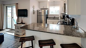 Une cuisine moderne dotée de plans de travail en granit, d’un four encastré et d’un réfrigérateur en acier inoxydable. On y trouve deux tabourets de bar et un téléviseur à écran plat fixé au mur.
