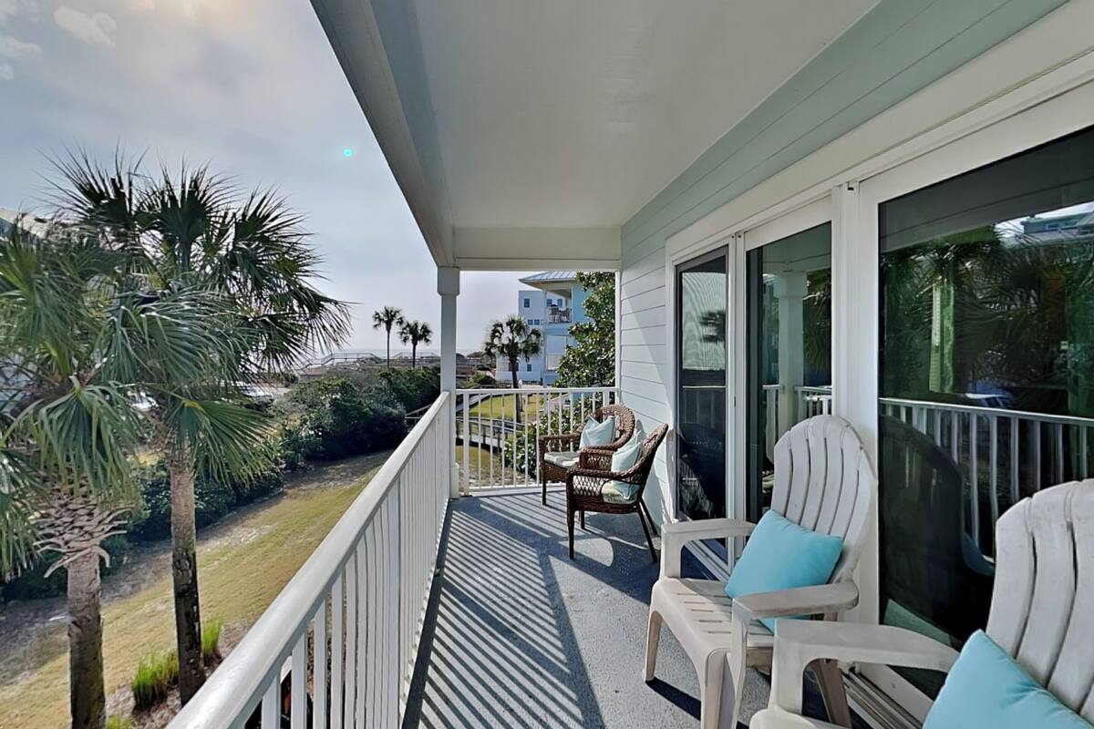 A balcony with white railings, two wicker chairs with cushions, and a view of palm trees and buildings.