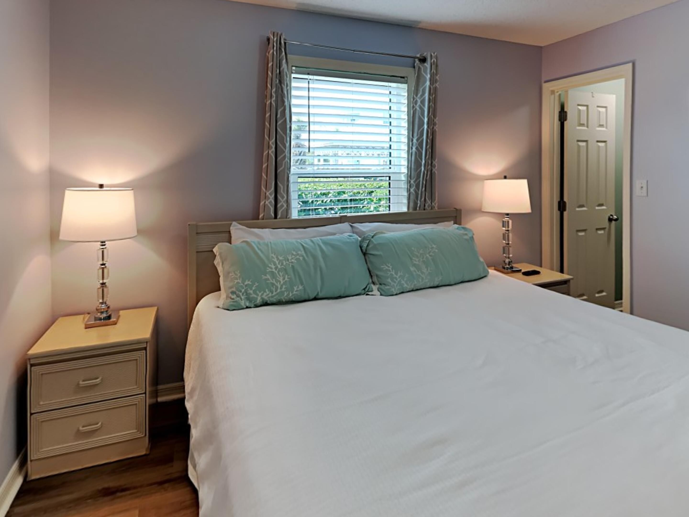 Une chambre à coucher comprenant un lit, deux tables de chevet, une commode, une fenêtre avec des stores et une porte.