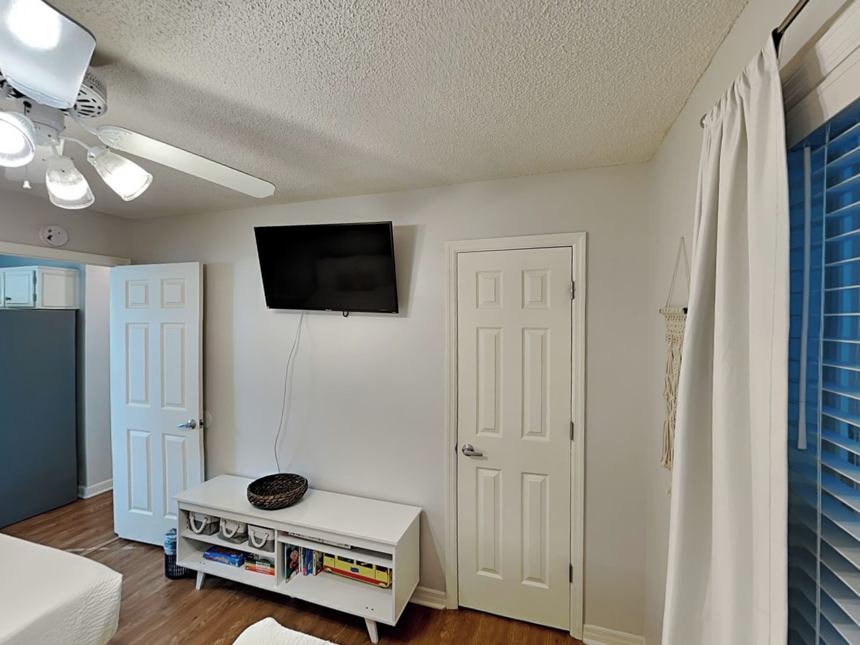 Un dormitorio con televisor montado en la pared, una puerta blanca, un armario blanco con espacio de almacenamiento y un ventilador de techo.