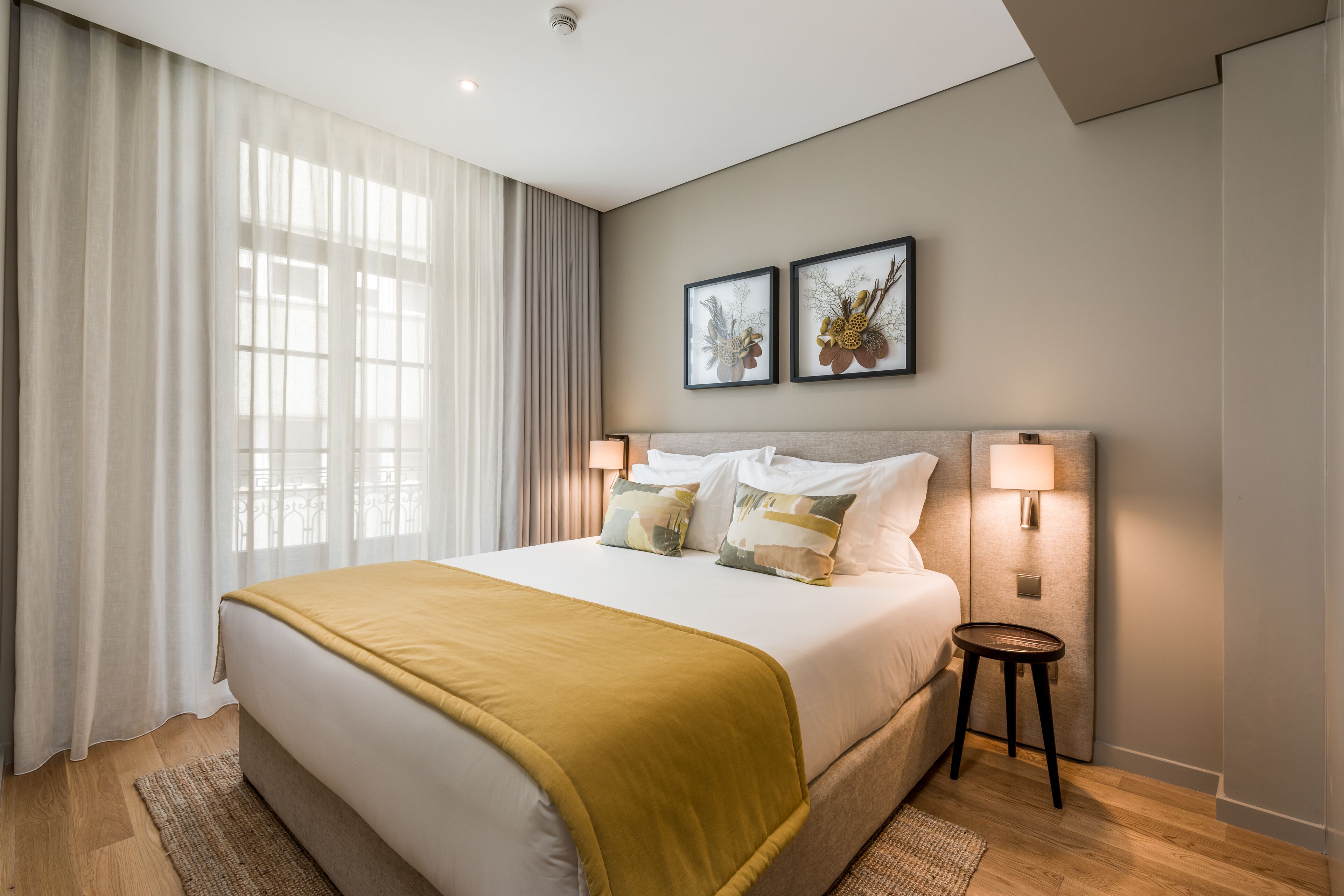 Una habitación de hotel moderna con una cama grande, dos lámparas de noche, una mesita auxiliar y un ventanal con cortinas translúcidas.