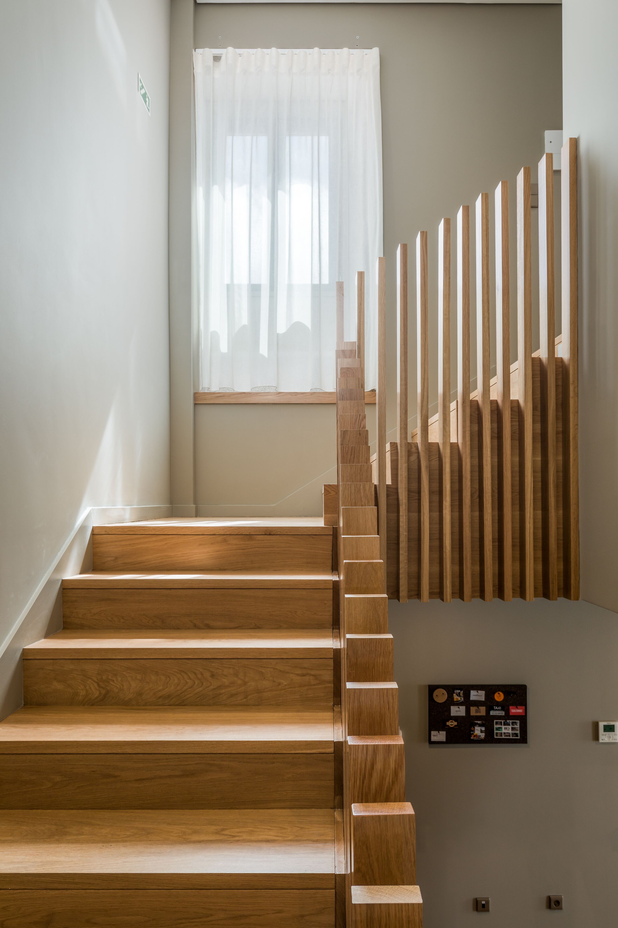A wooden staircase with a modern design, leading to a room with a window and a wall-mounted control panel.