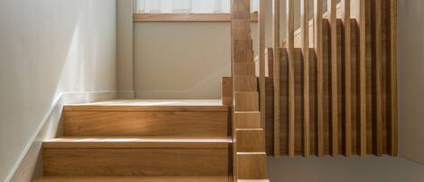 A wooden staircase with a modern design, leading to a room with a window and a wall-mounted control panel.