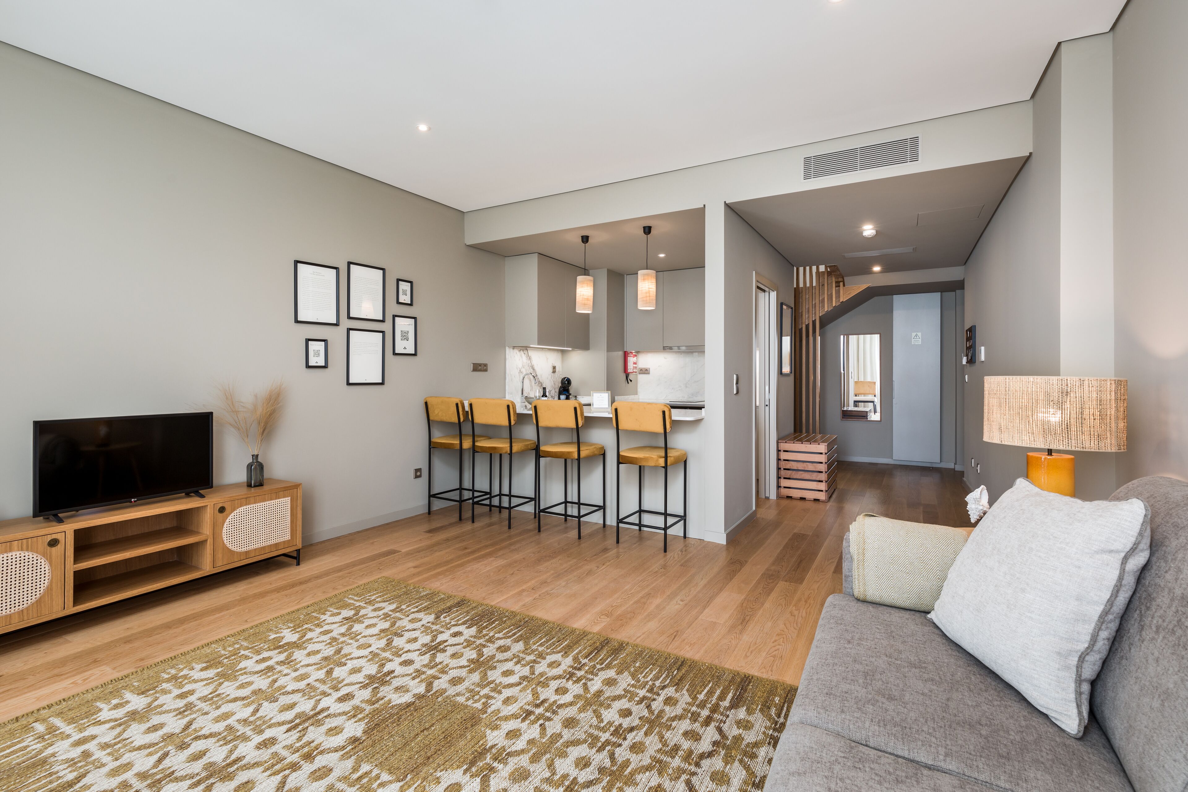 A modern living room with a flat-screen TV, a sofa, a patterned rug, and a kitchen area with bar stools.