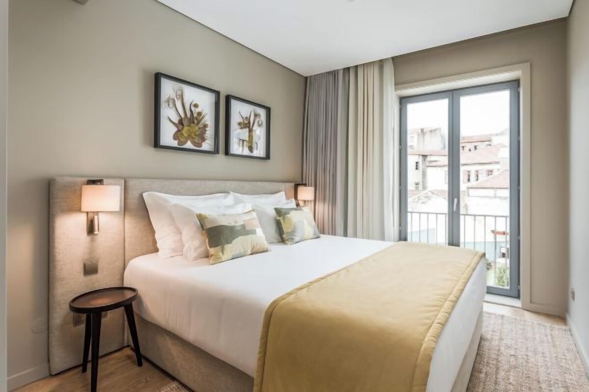 Une chambre d’hôtel avec un grand lit, deux lampes de chevet, une petite table d’appoint et un balcon donnant sur des immeubles.