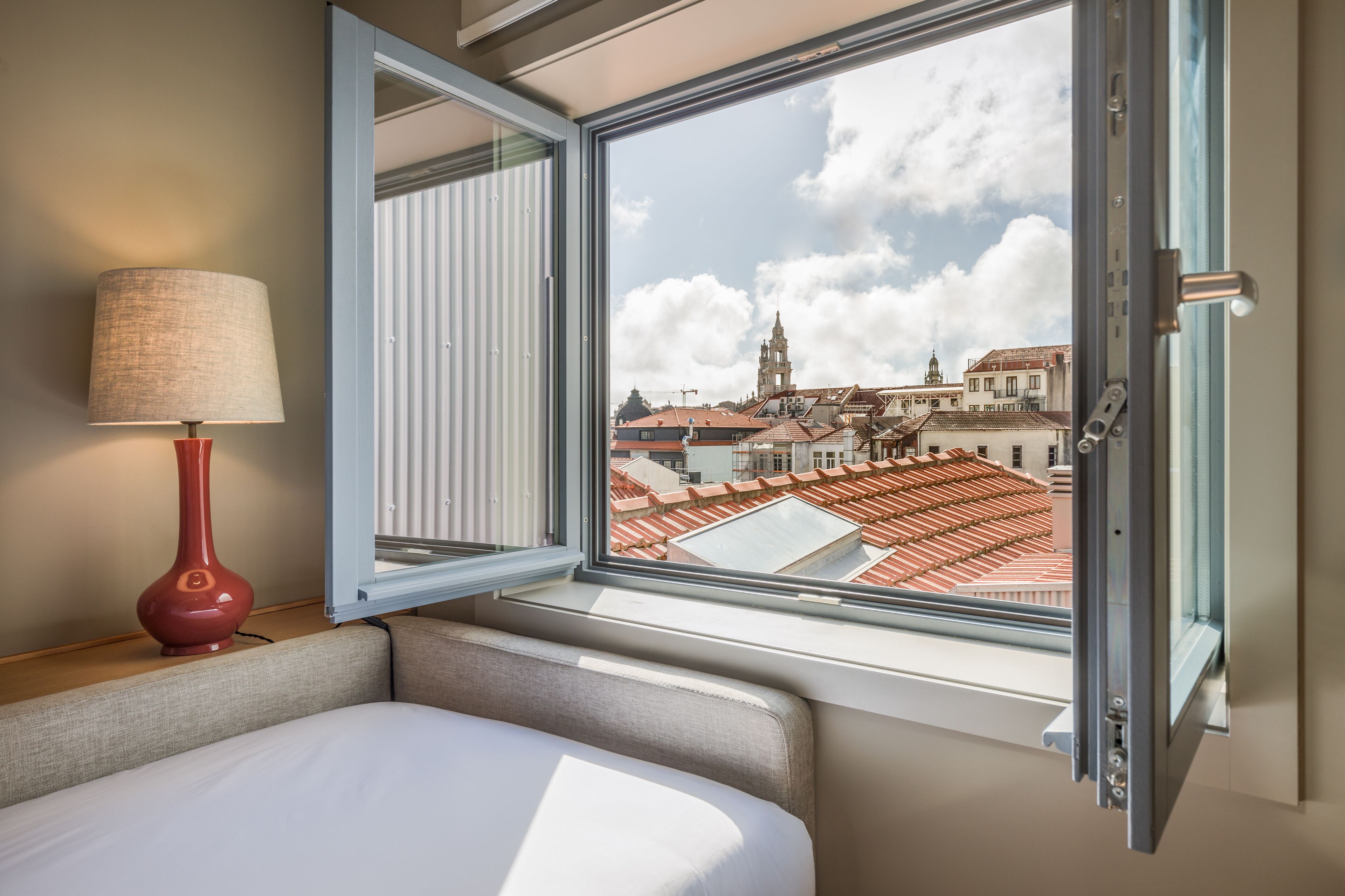 A hotel room with a bed, a window with a view of a cityscape, and a red lamp on the nightstand.