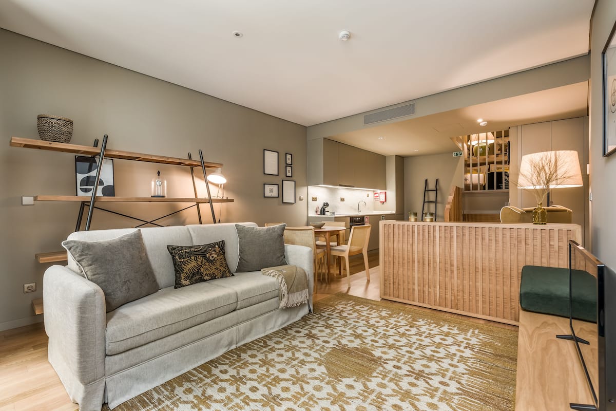 Ein modernes Wohnzimmer mit einer Couch, einem Couchtisch und einem gemusterten Teppich. Im Hintergrund befindet sich eine Küchenzeile mit Esstisch und Stühlen.