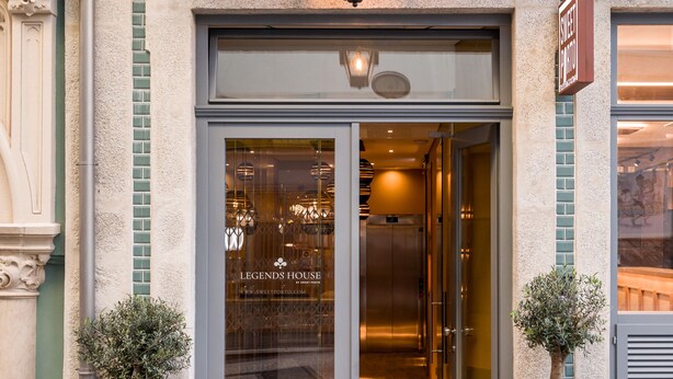 The entrance to a hotel with a glass door, a doormat, and two potted plants.
