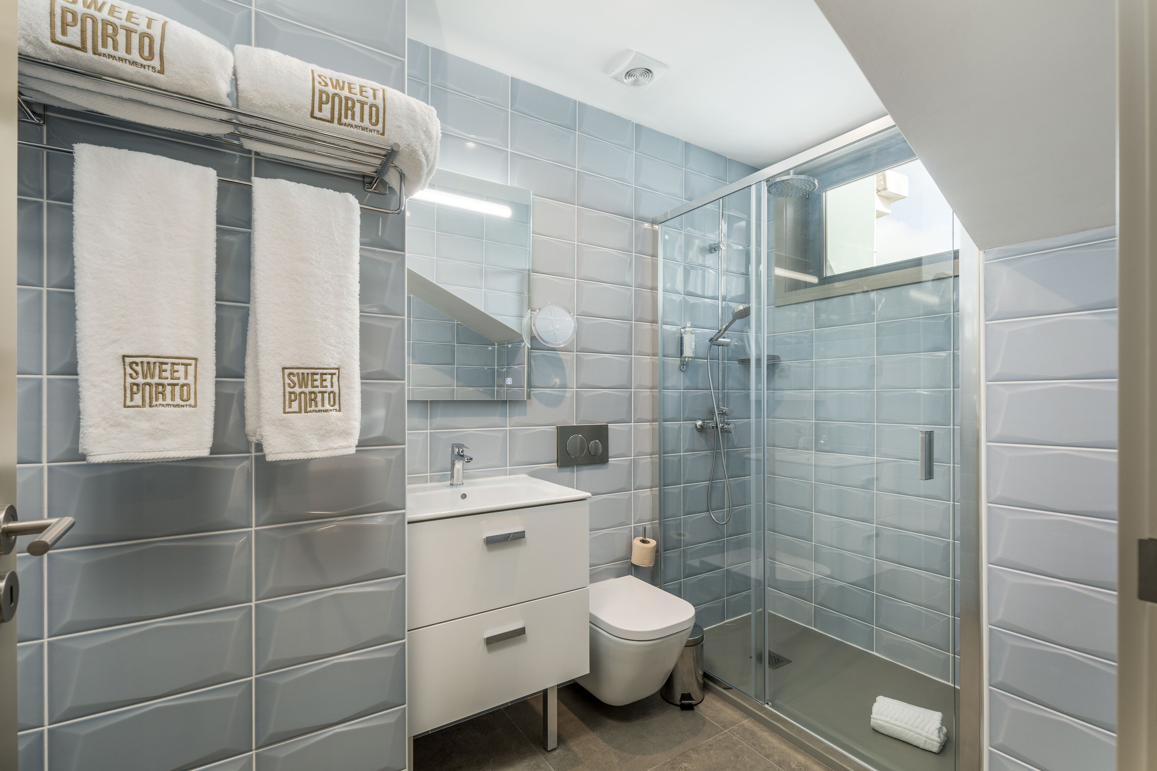 A bathroom with a glass shower enclosure, a white sink, and a toilet. There are towels hanging on the wall with the text "Sweet Porto Apartments".