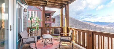 Una terraza de madera con mobiliario exterior, vistas a montañas y un edificio con fachada roja al fondo.