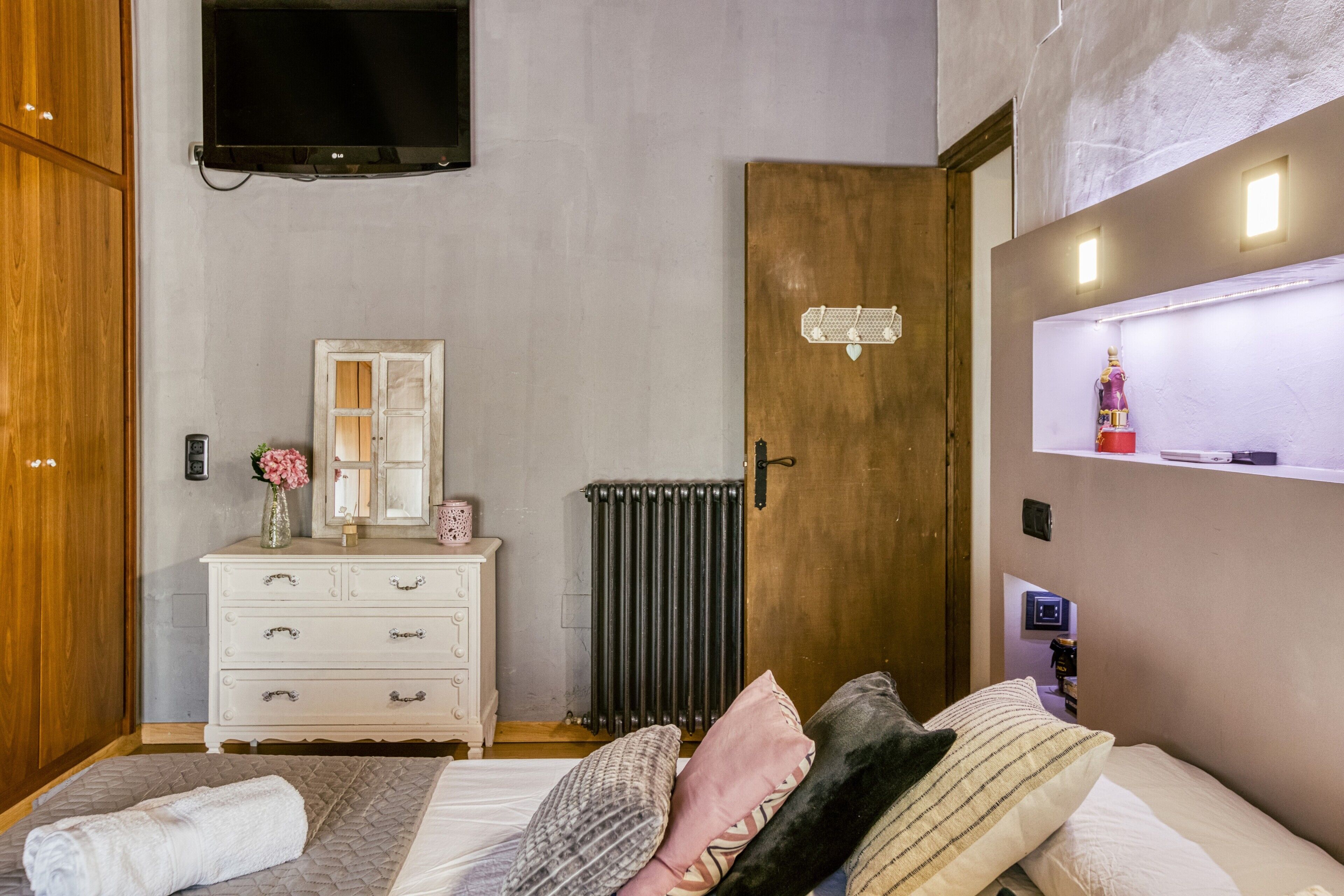 Ein Schlafzimmer mit einem Bett, einem Schminktisch, einem Spiegel, einer Heizung und einem an der Wand befestigten Fernseher.