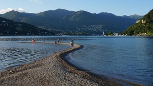 Une plage de galets où l’on voit des gens se promener le long du rivage, avec des montagnes et des bâtiments en arrière-plan.