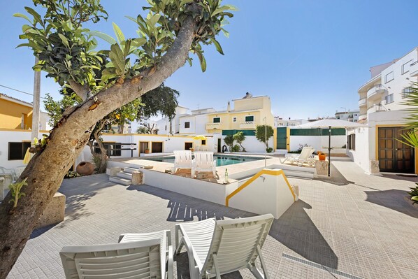 A pool area with white chairs, a tree, and a building with balconies.