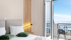 A hotel room with a bed, a nightstand, a wall-mounted lamp, a balcony with a chair, and a view of the sea.