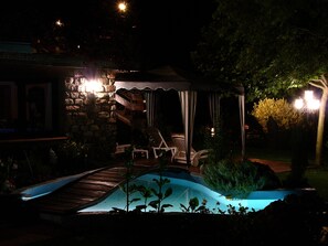 Un área de piscina exterior de noche con un deck de madera, sillas de descanso y una zona de estar cubierta.