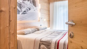 A bedroom with a bed, a wall-mounted light, a door with a handle, and a window with curtains. - Apartment Ciasa Rosa with Mountain View, Balconies & Shared Garden (Canazei)