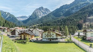 A mountain village with traditional houses, a central building with a balcony, and a road leading to a mountain pass. - Apartment Ciasa Rosa with Mountain View, Balconies & Shared Garden (Canazei)