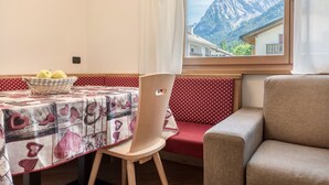 A dining area with a table set for two, a wicker basket with fruits, a red and white patterned tablecloth, a wooden chair, and a view of mountains through a window. - Apartment Ciasa Rosa with Mountain View, Balconies & Shared Garden (Canazei)