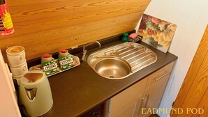 A compact kitchen with a stainless steel sink, a kettle, and a coffee maker. There are two bottles of LYONS juice on the counter.
