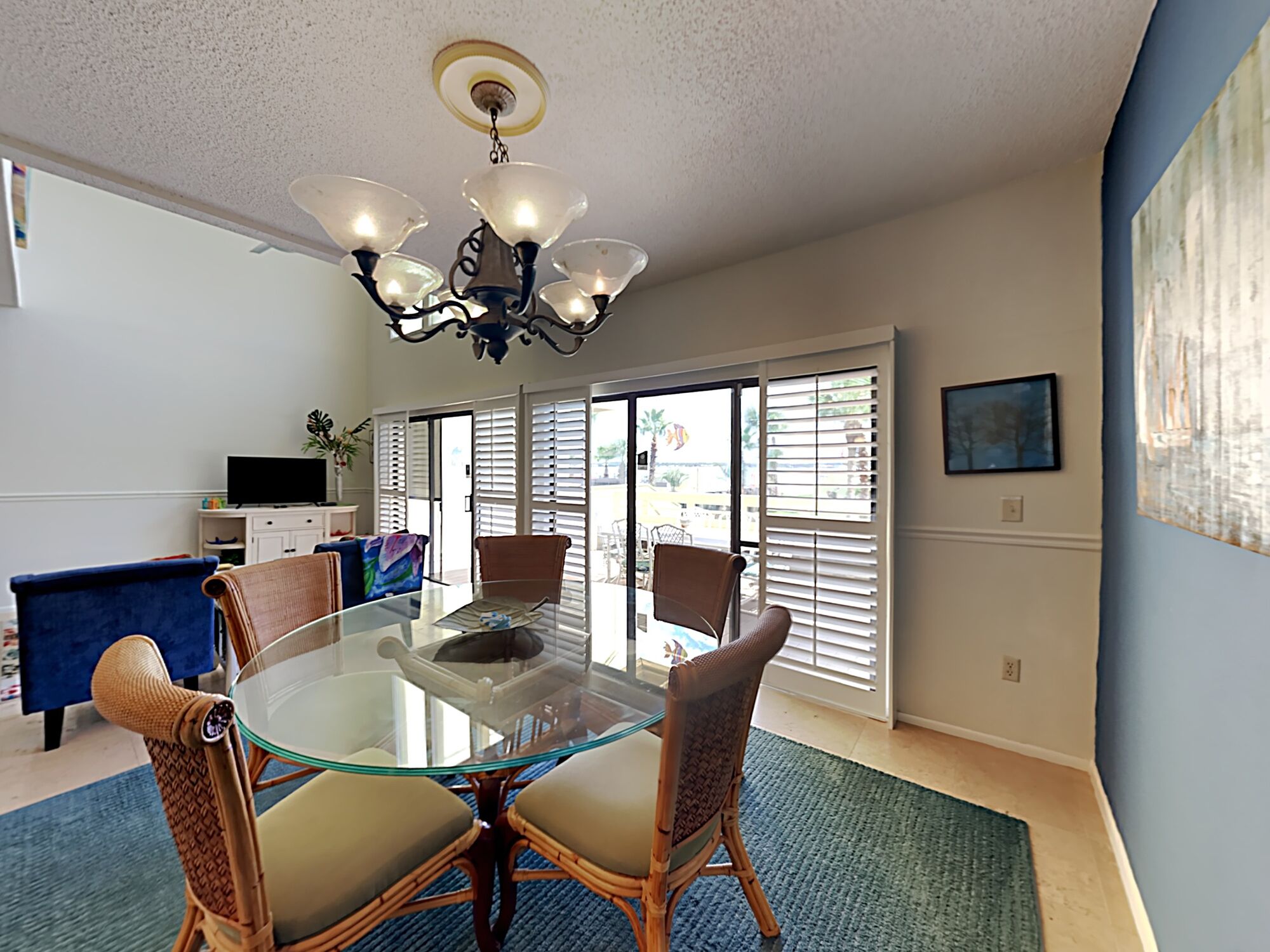 A dining area with a glass table, wicker chairs, a ceiling light fixture, and a view through large windows.