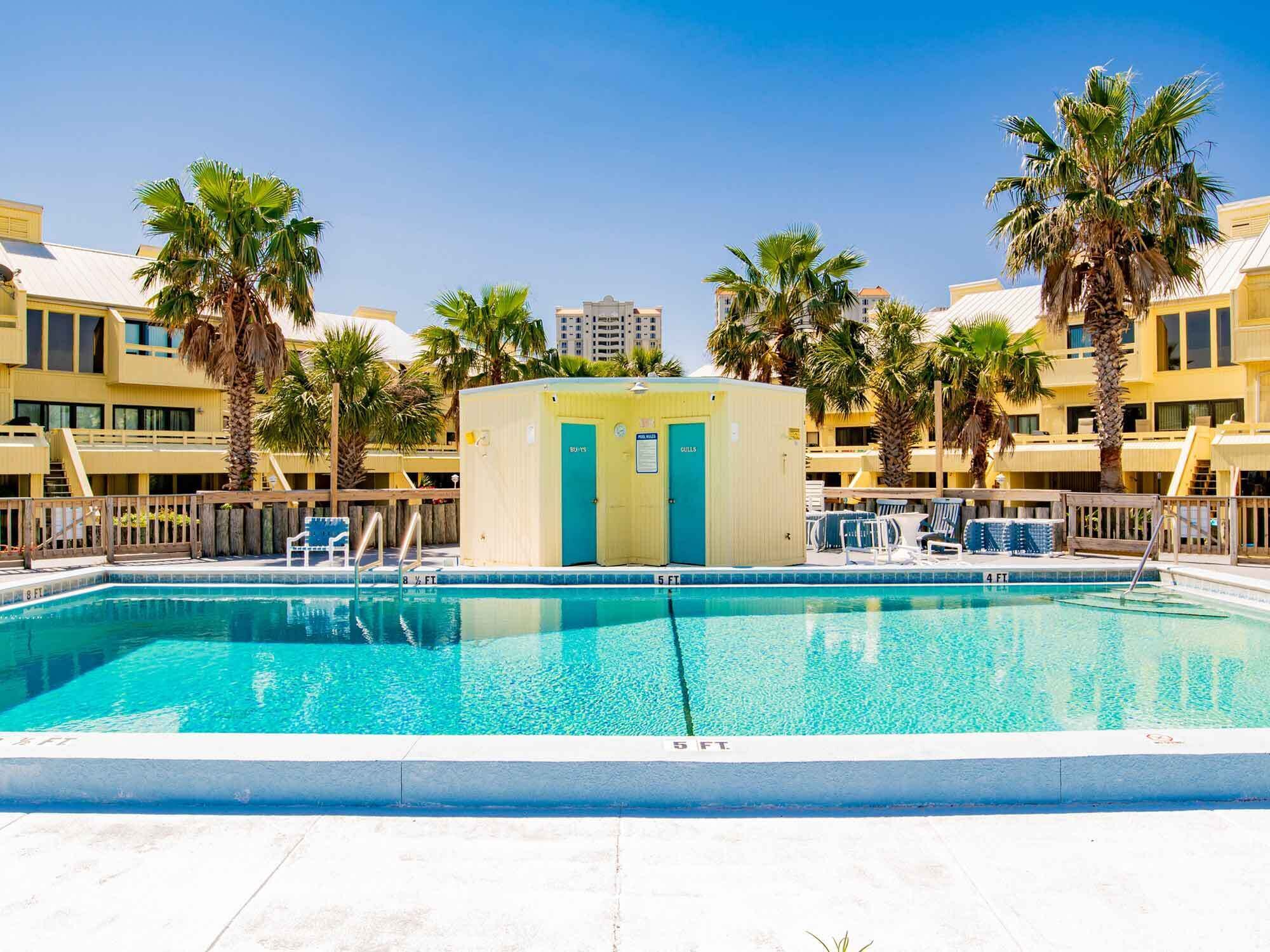 A swimming pool with a lifeguard station, palm trees, and a hotel building in the background.