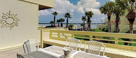 A deck with a table and chairs, overlooking a beach with palm trees and a clear sky.