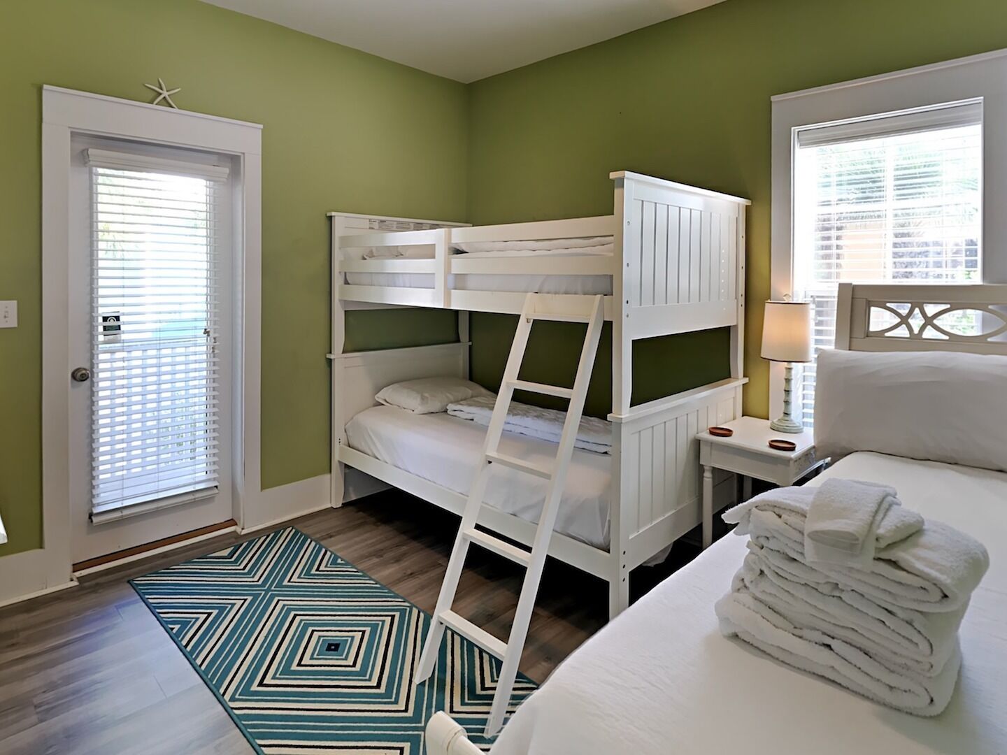 A bunk bed room with a door, a bedside table with a lamp, and a patterned rug.