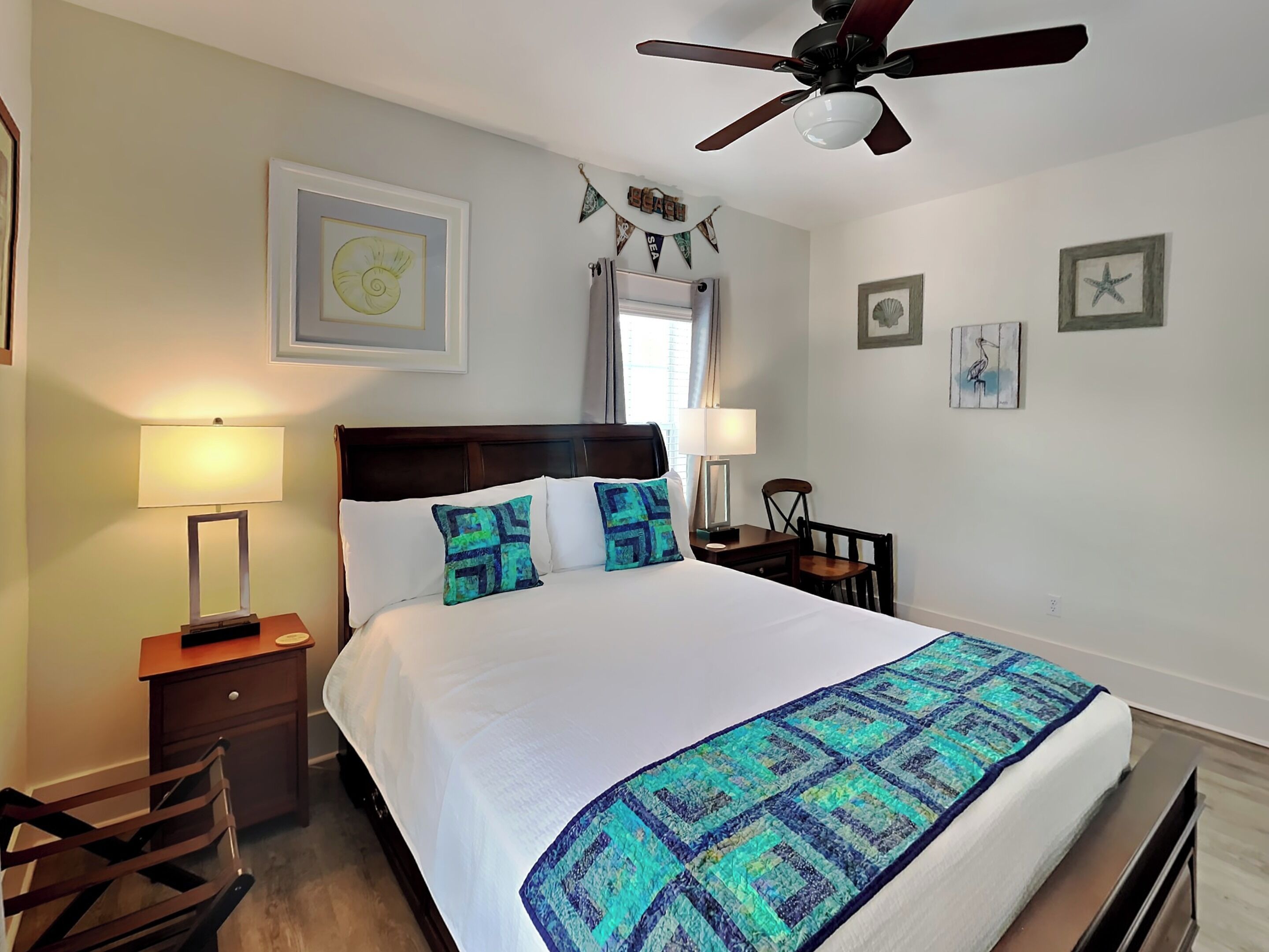 A neatly arranged bedroom with a bed, two bedside tables, a chair, a ceiling fan, and wall art.