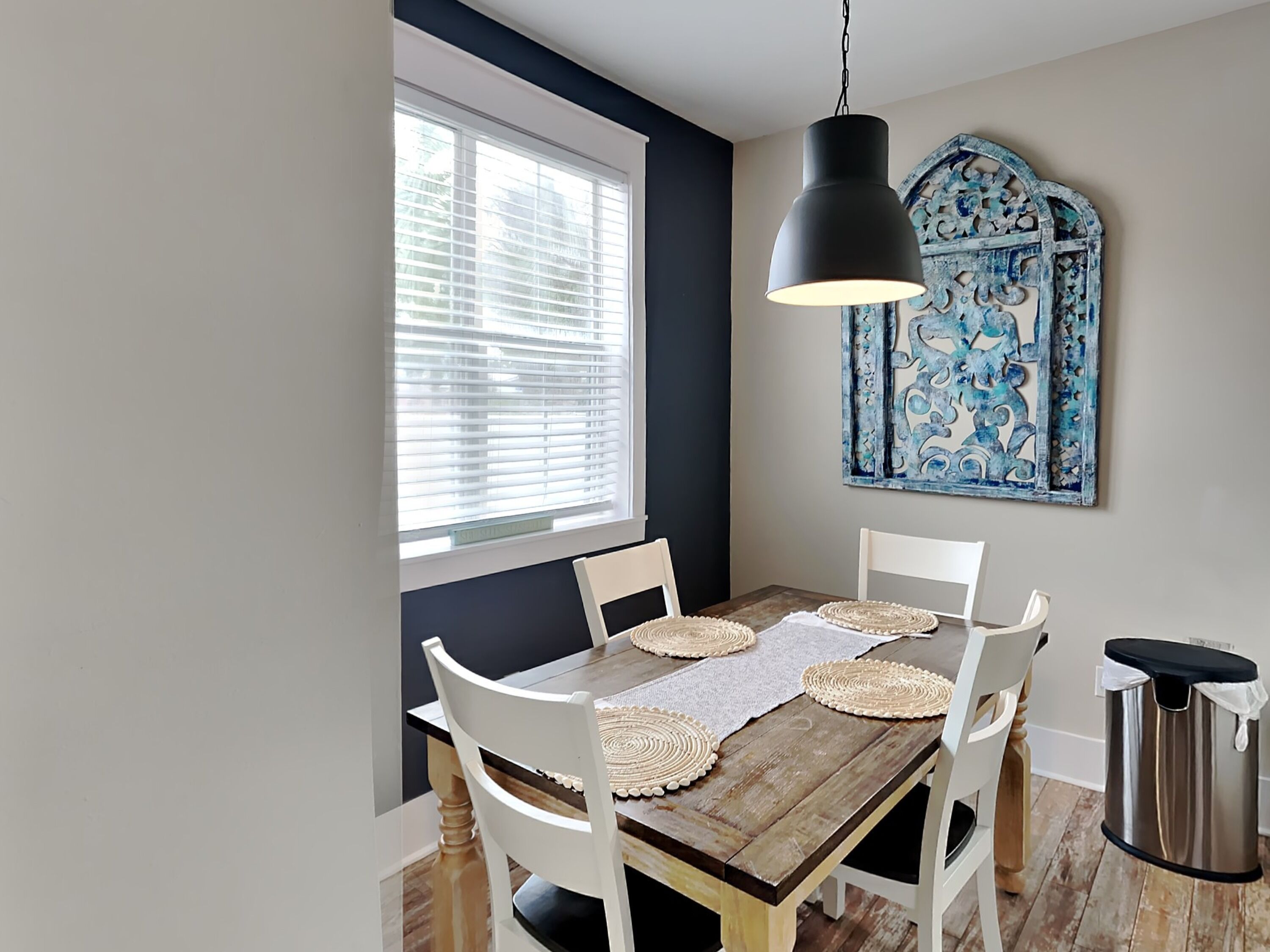 A dining area with a wooden table, white chairs, a black pendant light, and a decorative blue wall piece.