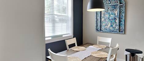 A dining area with a wooden table, white chairs, a black pendant light, and a decorative blue wall piece.