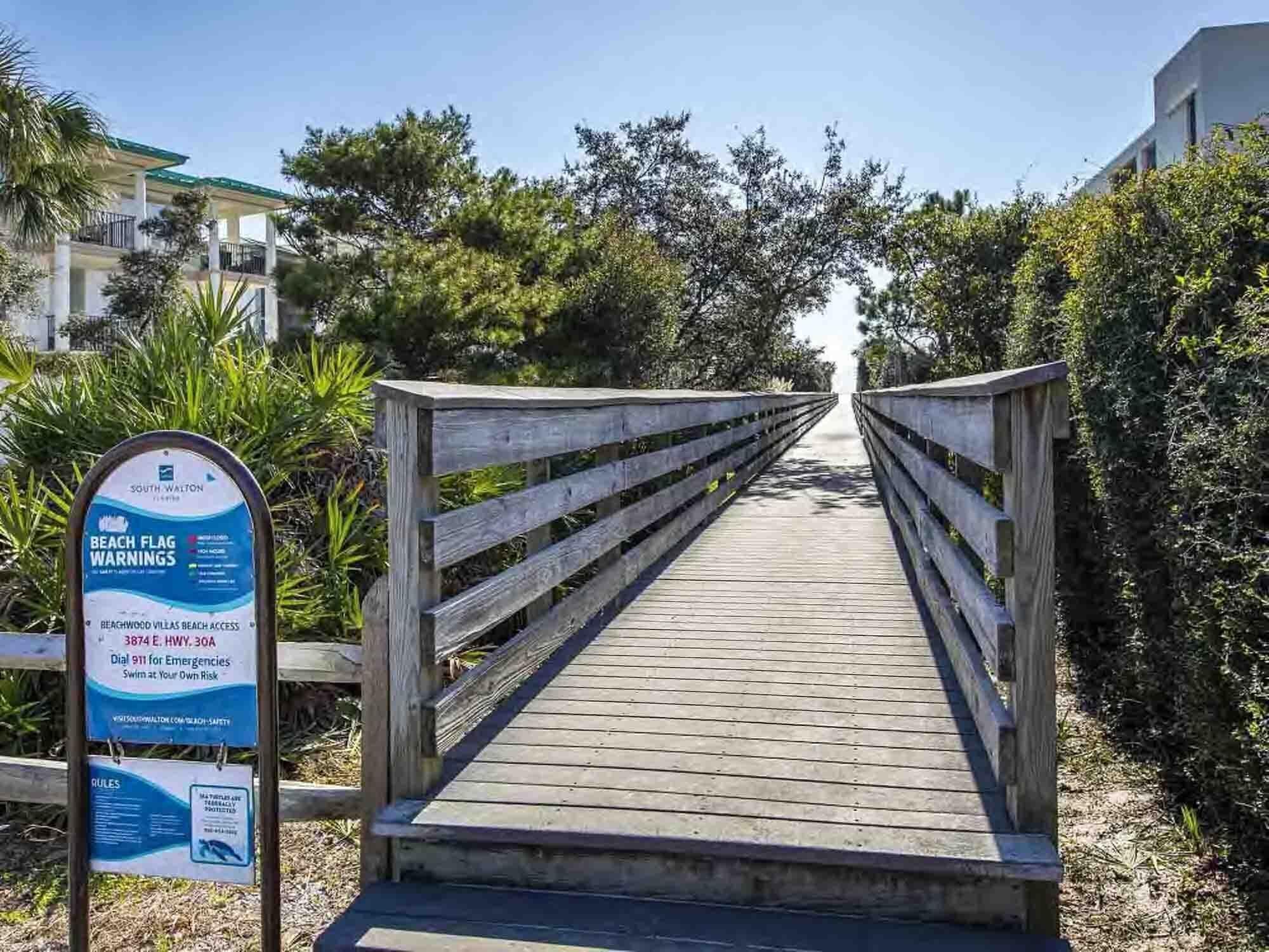 Une promenade en bois mène à l’accès à la plage de Beachwood Villas. Un panneau indique les consignes de sécurité concernant les drapeaux de signalisation sur la plage et fournit les coordonnées des personnes à contacter en cas d’urgence.