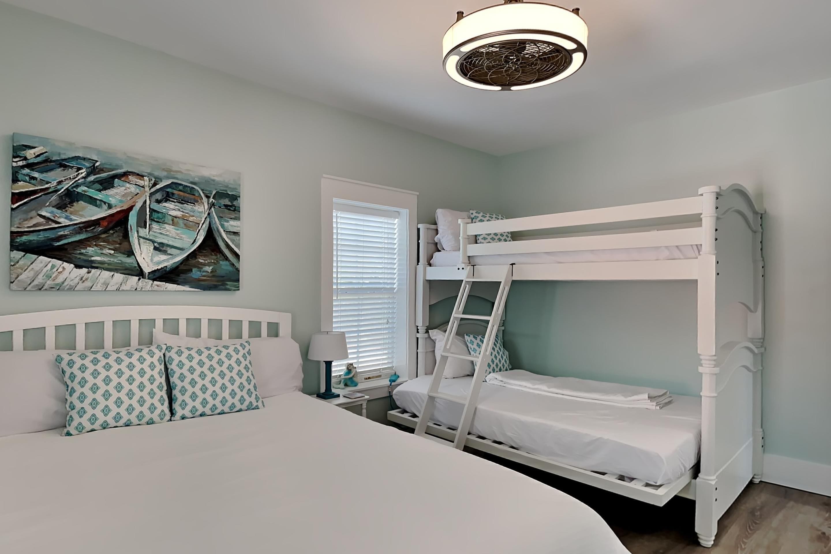 A bedroom with a bunk bed, a large bed, a painting of boats, a window with blinds, and a ceiling fan.