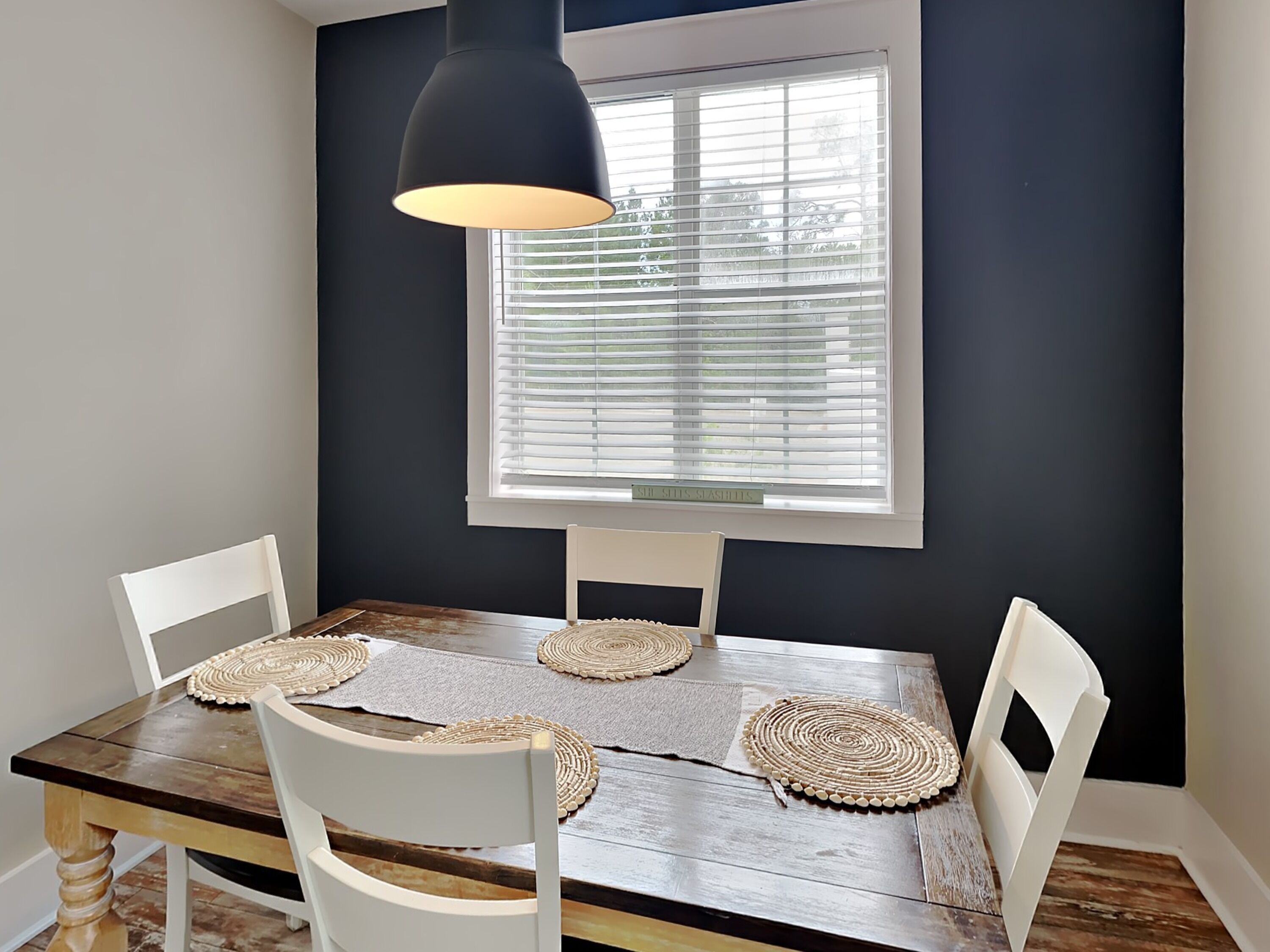 A dining area with a wooden table, white chairs, woven placemats, and a black pendant light.