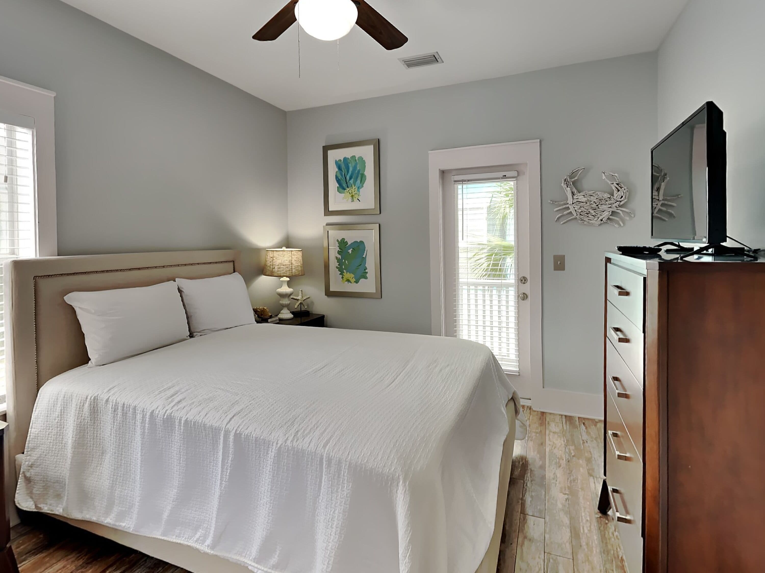 A bedroom with a large bed, a ceiling fan, a nightstand with a lamp, framed artwork on the wall, a door with a view, and a TV on a wooden cabinet.