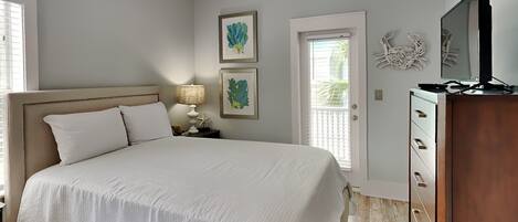A bedroom with a large bed, a ceiling fan, a nightstand with a lamp, framed artwork on the wall, a door with a view, and a TV on a wooden cabinet.