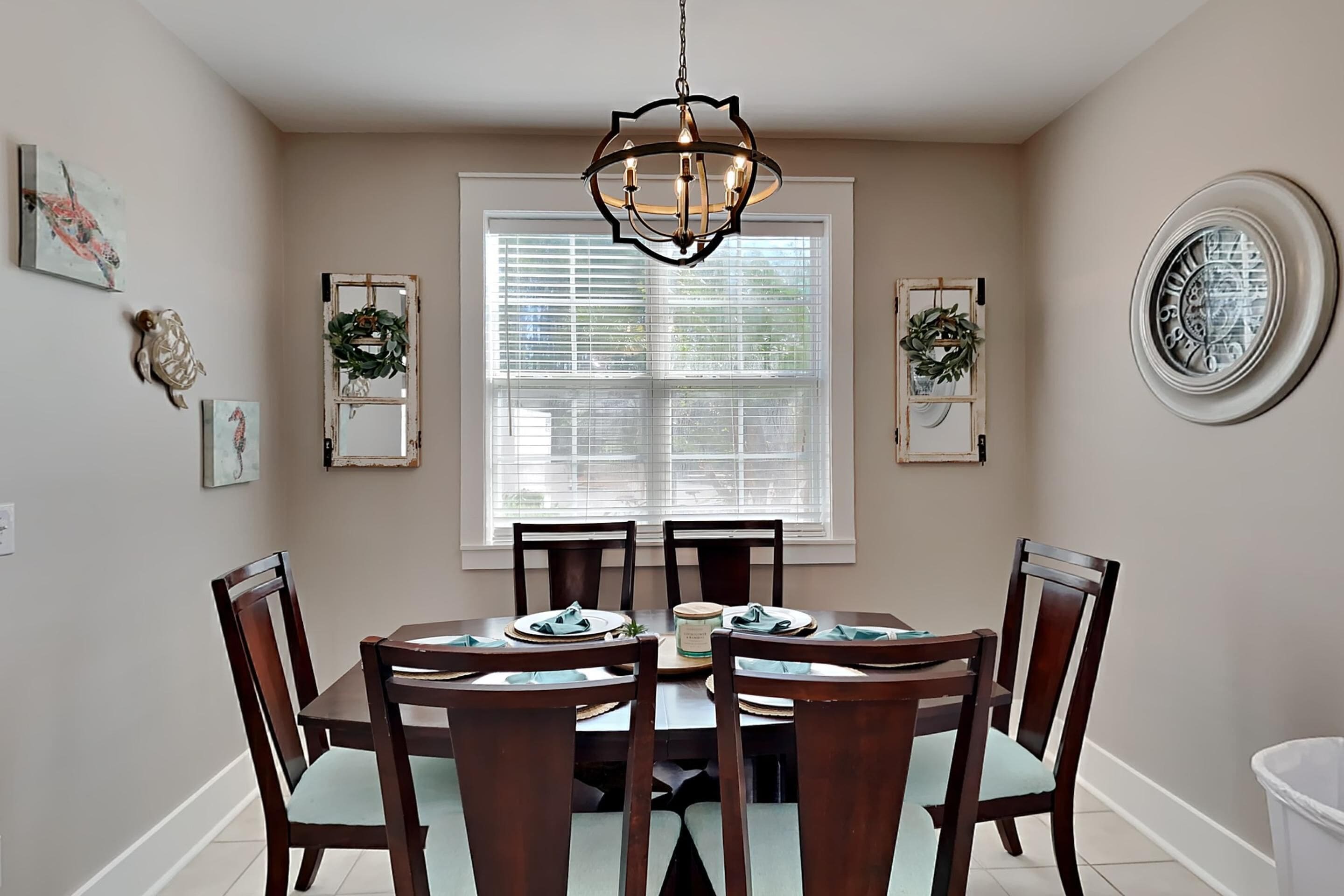 A dining room with a round table, six chairs, a chandelier, a window with blinds, and wall decorations.