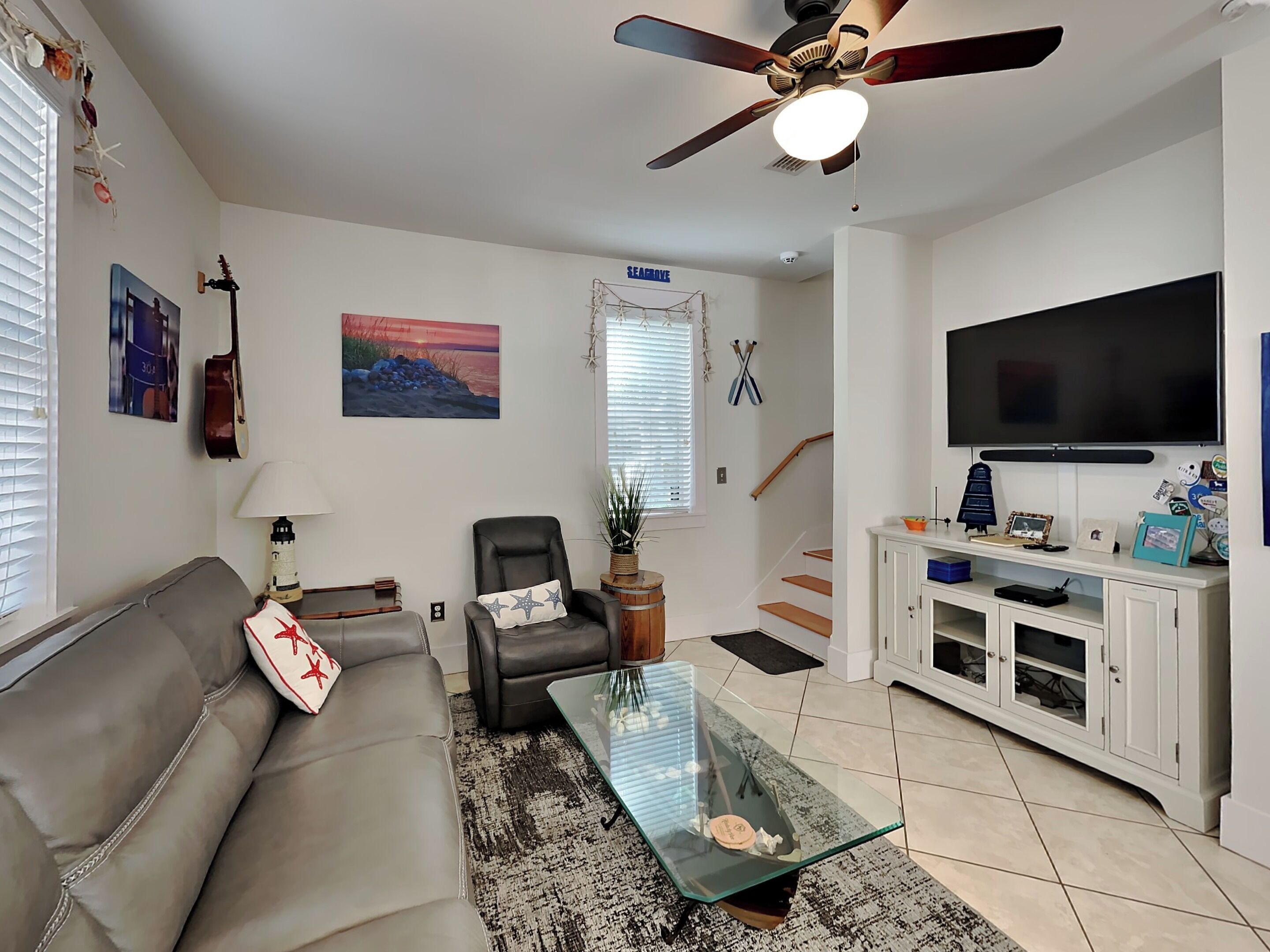 A living room with a sofa, a glass coffee table, a ceiling fan, a TV, and a staircase.